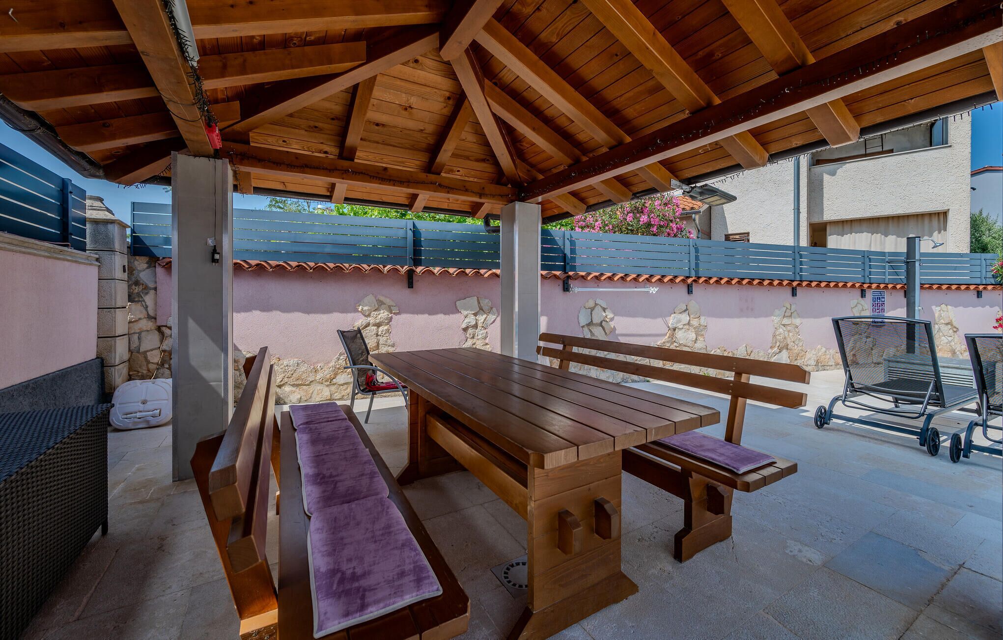 Een terras buiten met houten tafels en banken, stenen muren en een blauw hek.