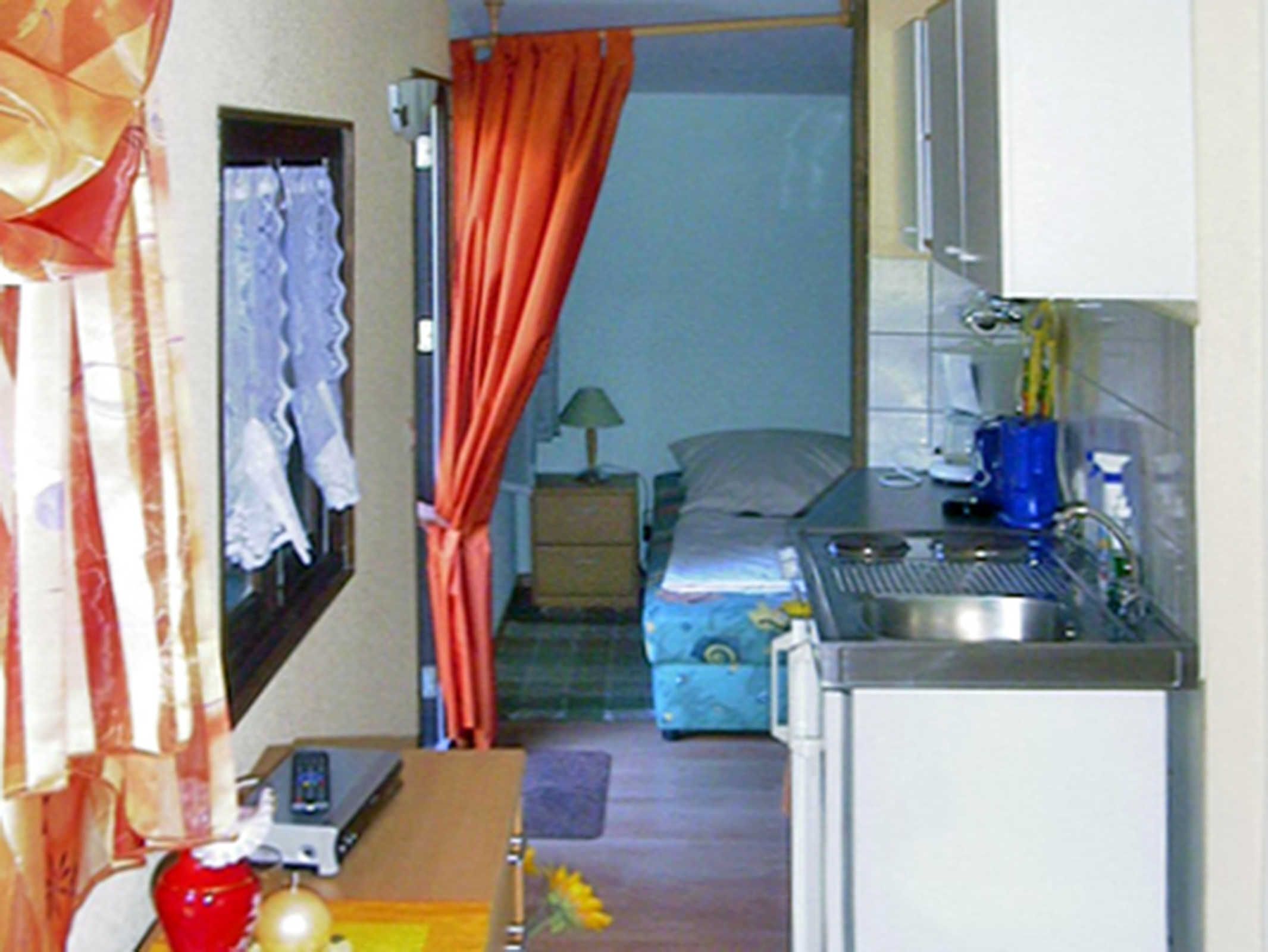 A compact hotel room with a kitchenette, a bed, a suitcase, and a window with curtains.