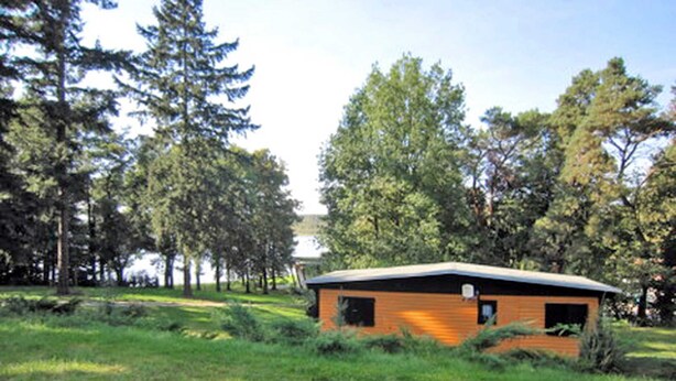 A small, single-story cabin with a sloped roof, surrounded by dense trees and grassy terrain.