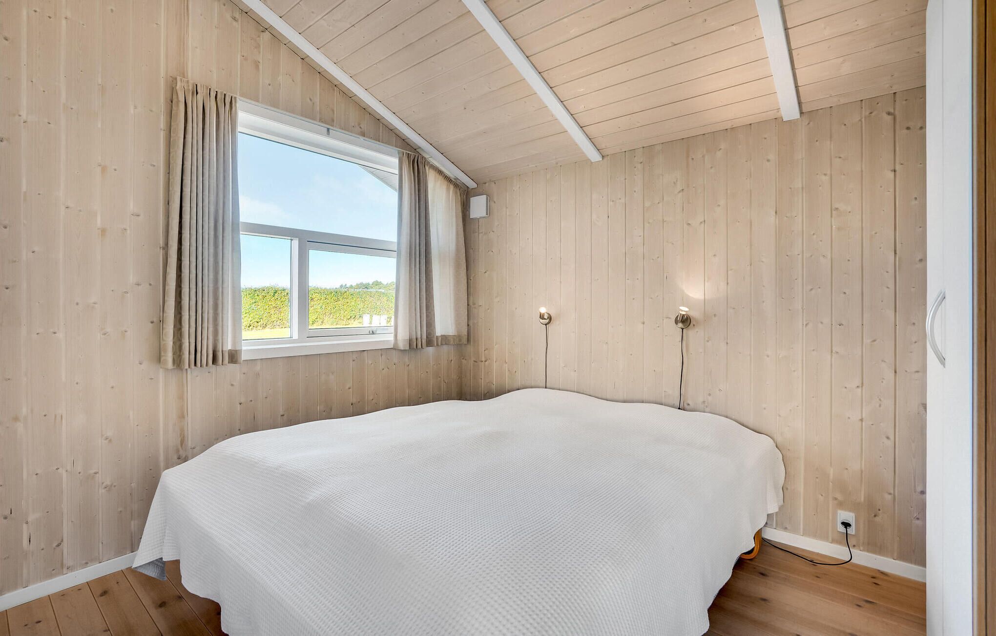 A bedroom with a large bed, wooden walls, a window with curtains, and a view of greenery outside.