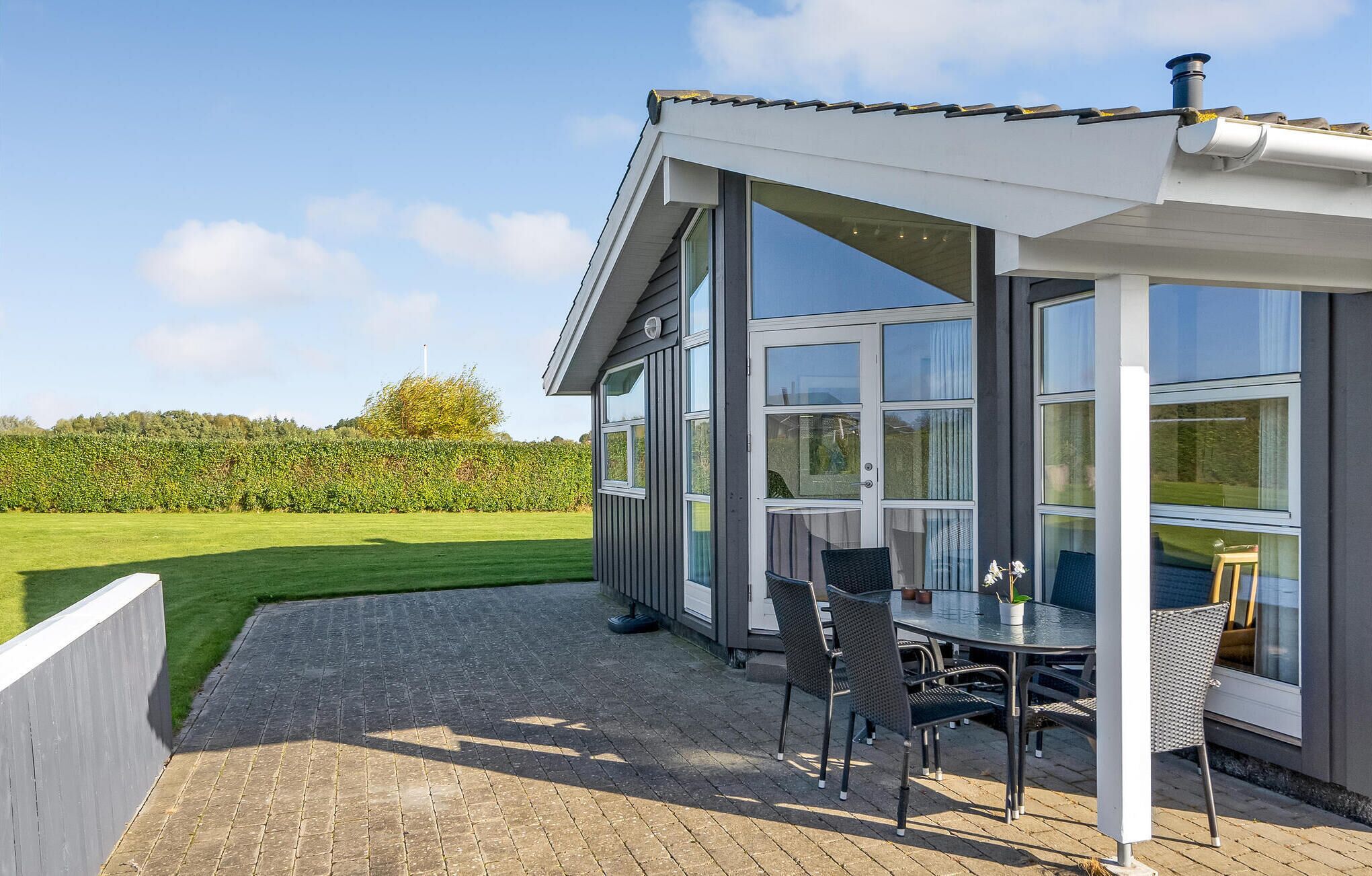 Un patio exterior moderno con mesa y sillas, ventanas amplias y vista a un campo verde.