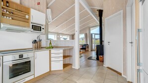 Eine moderne Küche mit weißen Schränken, einem Einbauofen und einer Mikrowelle. Ein Holzregal beherbergt Gläser und eine Weinflasche. Die Küche geht in einen Essbereich mit einem schwarzen Grill und Blick ins Freie über.