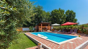 Une piscine avec un transat et un parasol rouge, entourée de verdure et d’une terrasse en briques.