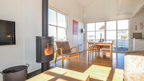 Une pièce chaleureuse avec un poêle à bois, un coin repas avec des meubles en bois et de grandes fenêtres donnant sur l’extérieur.
