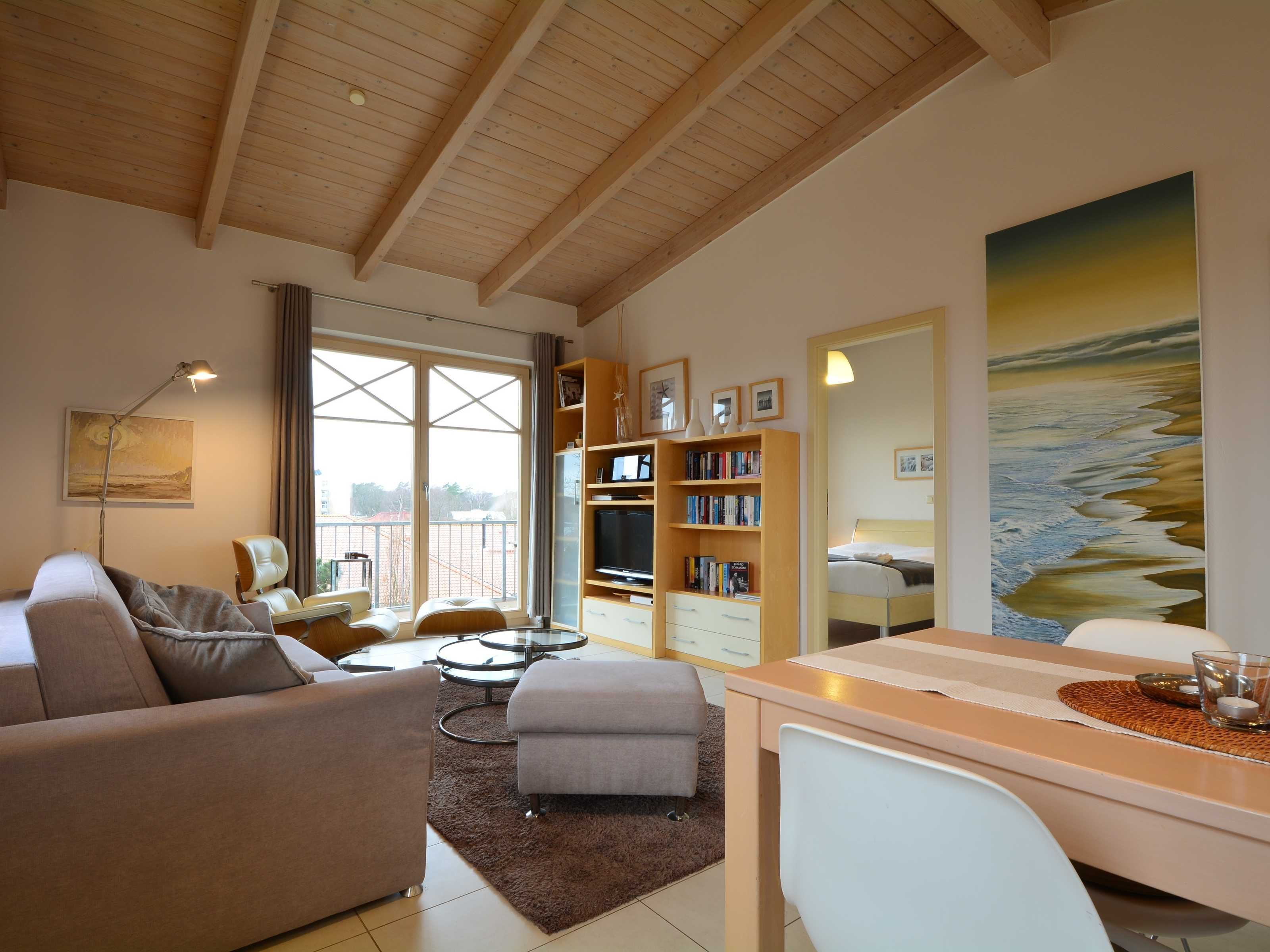 Un salon spacieux avec un plafond haut, une grande fenêtre, un canapé, une table basse, une bibliothèque et un tableau représentant une scène de plage.