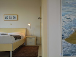 Quarto com cama, mesa de cabeceira, luminária e uma pintura de paisagem marítima.