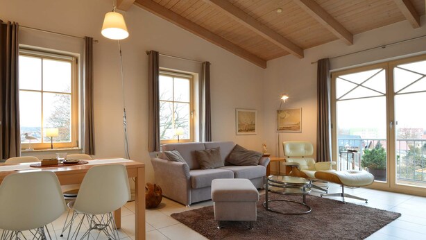 A spacious living room with a wooden ceiling, large windows, a grey sofa, a dining table with chairs, and a small round table with a chair. - Residence Seestern Whg S-13. - See-13 (Kühlungsborn)