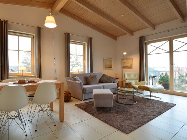 Un salon spacieux avec un plafond en bois, de grandes fenêtres, un canapé gris, une table à manger avec des chaises et une petite table ronde avec un siège.