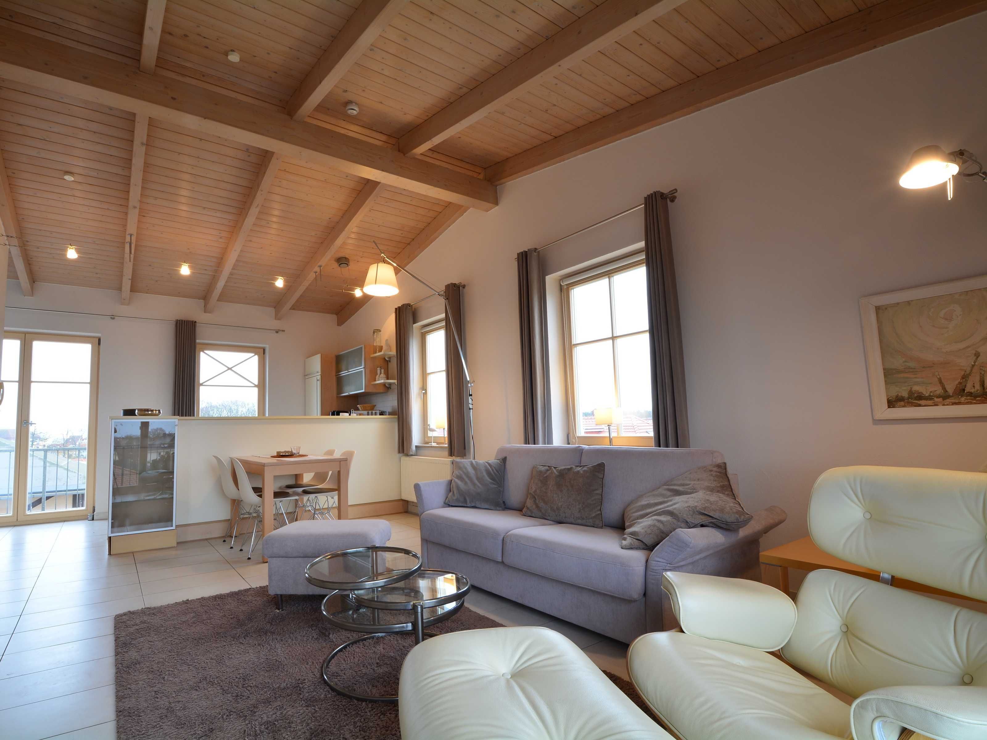Un salón espacioso con techo alto, un sofá gris, un sillón blanco, una mesa de centro de vidrio y una cocina al fondo.