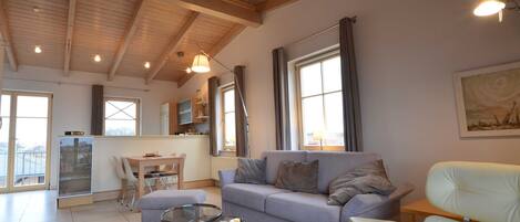 A spacious living area with a high ceiling, a grey sofa, a white armchair, a glass coffee table, and a kitchen in the background.