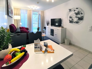Ein modernes Wohnzimmer mit einer Couch, einem Esstisch mit Obst, einem Fernseher und einer Wanddekoration.