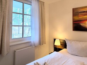 Ein Hotelzimmer mit großem Fenster, Heizkörper, Nachttischlampe, einem Bett mit Kissen und einem Bild an der Wand.