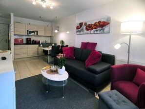 Un salon moderne avec un canapé noir, des coussins rouges, une table basse agrémentée d’objets décoratifs, un coin repas avec une chaise noire et une cuisine équipée d’un four à micro-ondes.