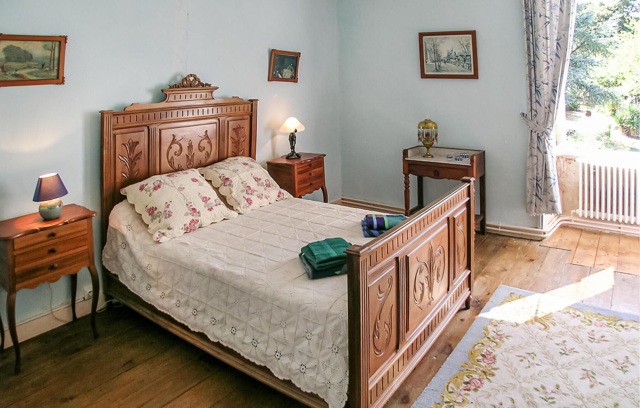 Ein Schlafzimmer mit einem hölzernen Bett, einem Nachttisch mit Lampe, einem kleinen Tisch mit Dekoration und einem Fenster mit Blick ins Grüne.