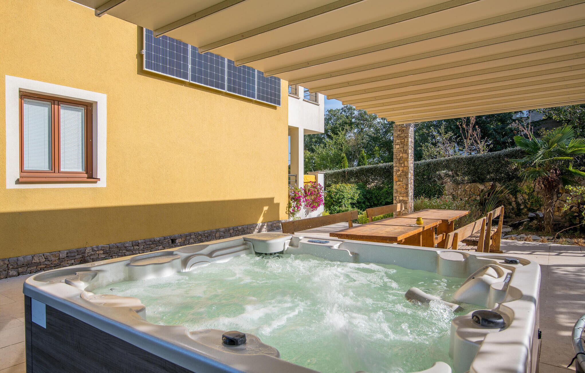 Ein Whirlpool unter einer überdachten Terrasse, mit einer gelben Wand, einem Fenster und Solaranlagen.