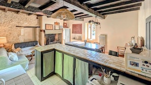 A rustic kitchen with a stone wall, a wooden ceiling, and a tiled island with checkered curtains. - 2 bedroom stunning home in Grignols (Grignols)