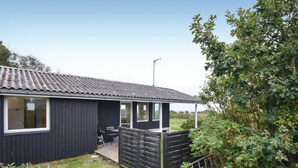 Un petit bâtiment de couleur sombre, doté d’un toit en tuiles, d’une terrasse en bois et d’un jardin clos.