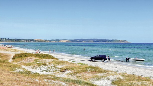 Une plage avec une voiture et un bateau sur le rivage, des gens dans l’eau et un ciel dégagé.