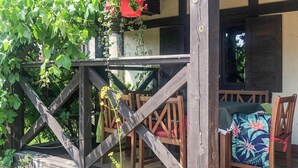 Eine hölzerne Veranda mit Bank und Tisch, umgeben von viel Grün und einem roten, hängenden Strauch.