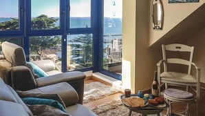 Ein gemütliches Wohnzimmer mit großem Fenster und schöner Aussicht, ein bequemes Sofa, ein kleiner Tisch mit Snacks und ein Sessel.