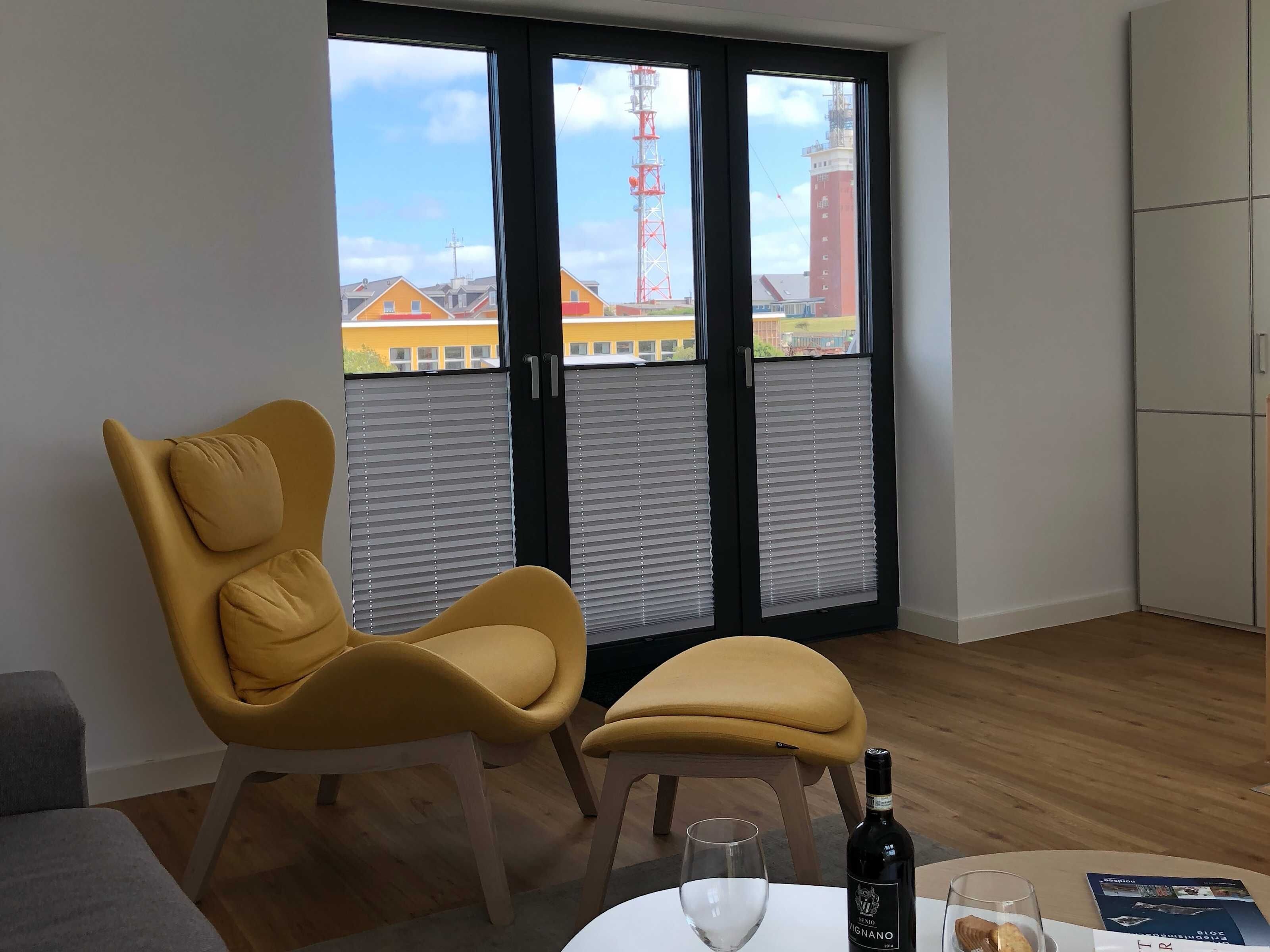Et værelse med en gul stol, et lille bord med en flaske og et glas, og udsigt til bygninger gennem et vindue.