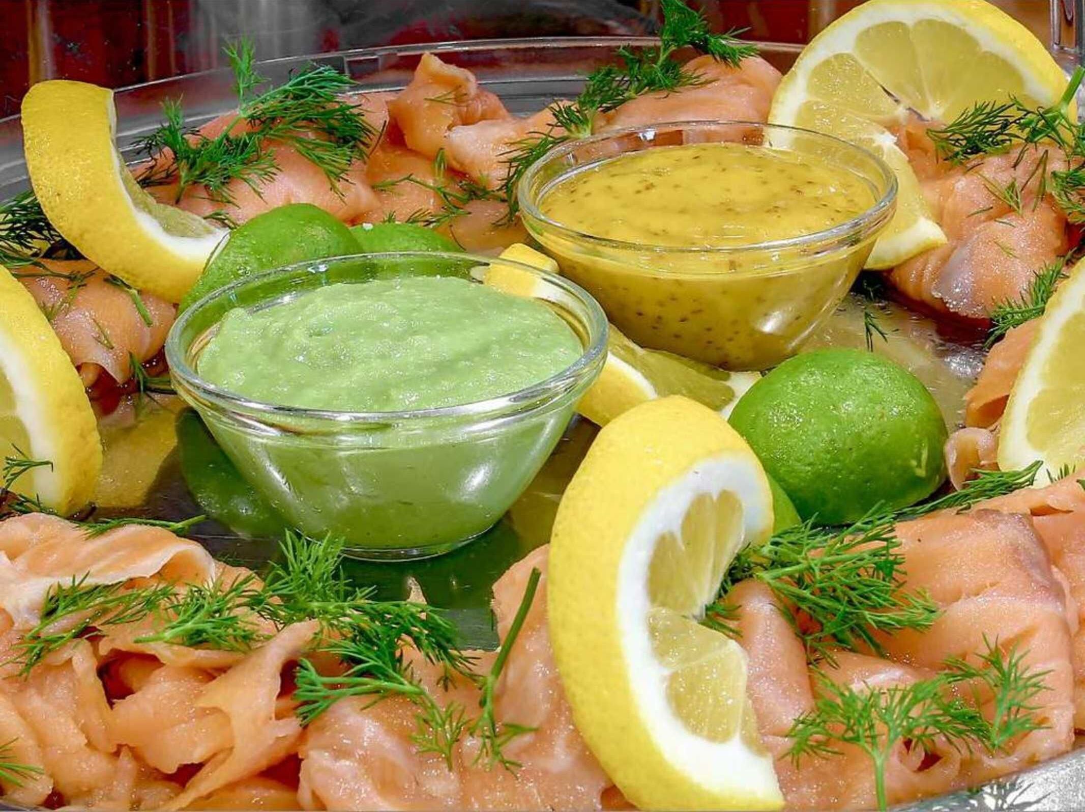 A platter of smoked salmon with lemon slices, dill, and two bowls of sauce.