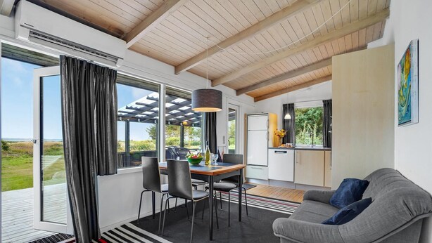 Una sala de estar moderna con un sofá gris, una mesa de comedor con sillas, una zona de cocina y ventanas grandes con vistas al exterior.