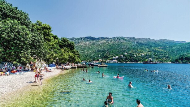 Una playa con agua cristalina, gente nadando y tomando el sol, barcos y montañas al fondo.