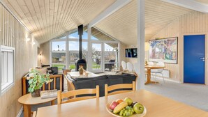 Un intérieur moderne avec un plafond en bois, une cheminée, un coin repas agrémenté d’un panier de fruits et une grande fenêtre donnant sur l’extérieur.