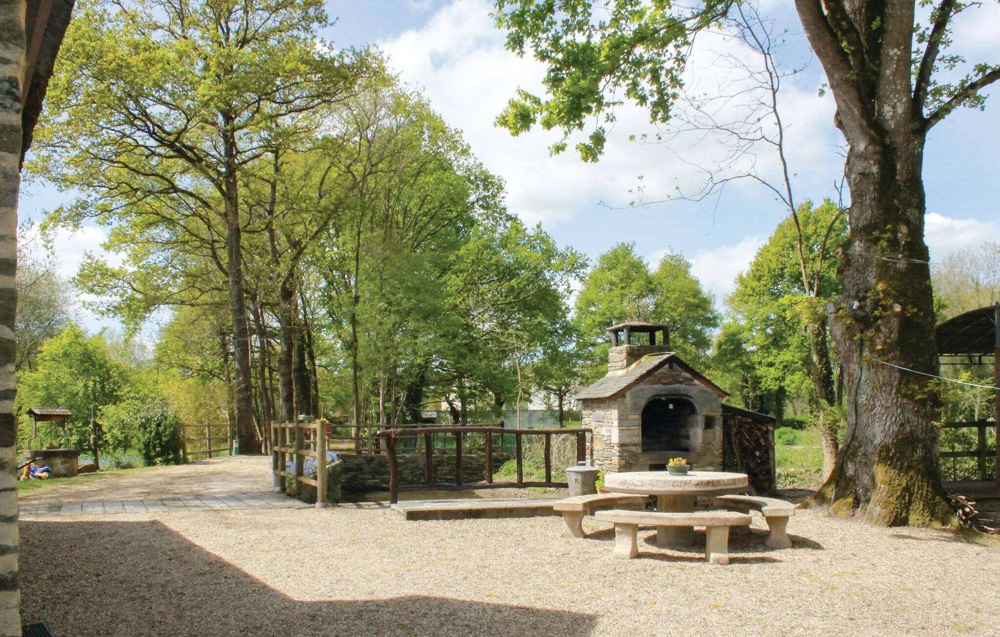 A rustic outdoor area with a stone-built structure, possibly a barbecue or grill, surrounded by trees and a gravel path.