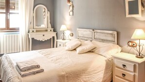 Een slaapkamer met een groot bed, witte beddengoed, een dressoir met lades, een nachtkastje met een lamp, een spiegel en een fotolijst aan de muur.