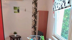 Ett litet rum med trägolv, ett litet matbord med en stol, ett fönster med utsikt över grönområde, en liten pall med en växt och en säng med ett rött och vitt sängöverkast.