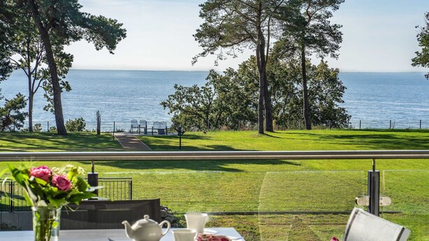 A table with a vase of flowers, teapot, cups, and saucers, overlooking a grassy area with trees and a view of the sea. - Apartment Haffperle - Oasis on the lagoon - Haffperle (1-4) (Garz/Usedom)
