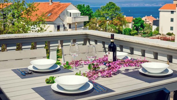 Une table dressée pour deux personnes, avec de l’argenterie blanche, des verres à vin et une bouteille, sur un balcon donnant sur une ville côtière.