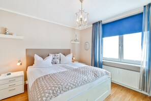Une chambre à coucher bien aménagée, avec un lit, des tables de chevet, une commode, un lustre, une fenêtre avec des rideaux et une étagère murale décorée de divers objets.
