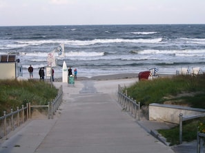Ein Küstenweg führt zum Strand, wo ein Schild 'Streer kust' und ein Rettungsschwimmerstand zu sehen sind.