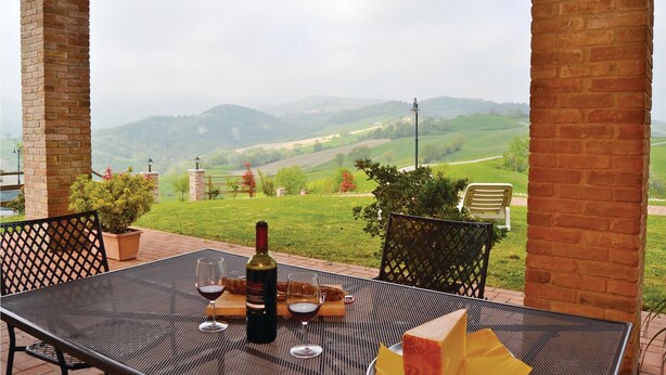 Une table de patio avec une bouteille de vin, des verres et une planche en bois garnie de pain, le tout placé devant un paysage pittoresque de collines ondulantes et d’un ciel nuageux.