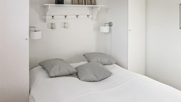 A minimalist bedroom with a white bed, striped pillows, a shelf with decorative items, and a wall-mounted lamp.