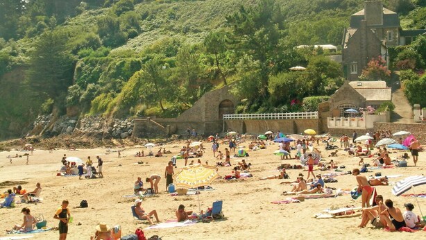 Ein überfüllter Strand mit Menschen, die sich sonnen und schwimmen. Es gibt Sonnenschirme und Liegestühle. Im Hintergrund ist ein Gebäude mit einem steilen Dach zu sehen.