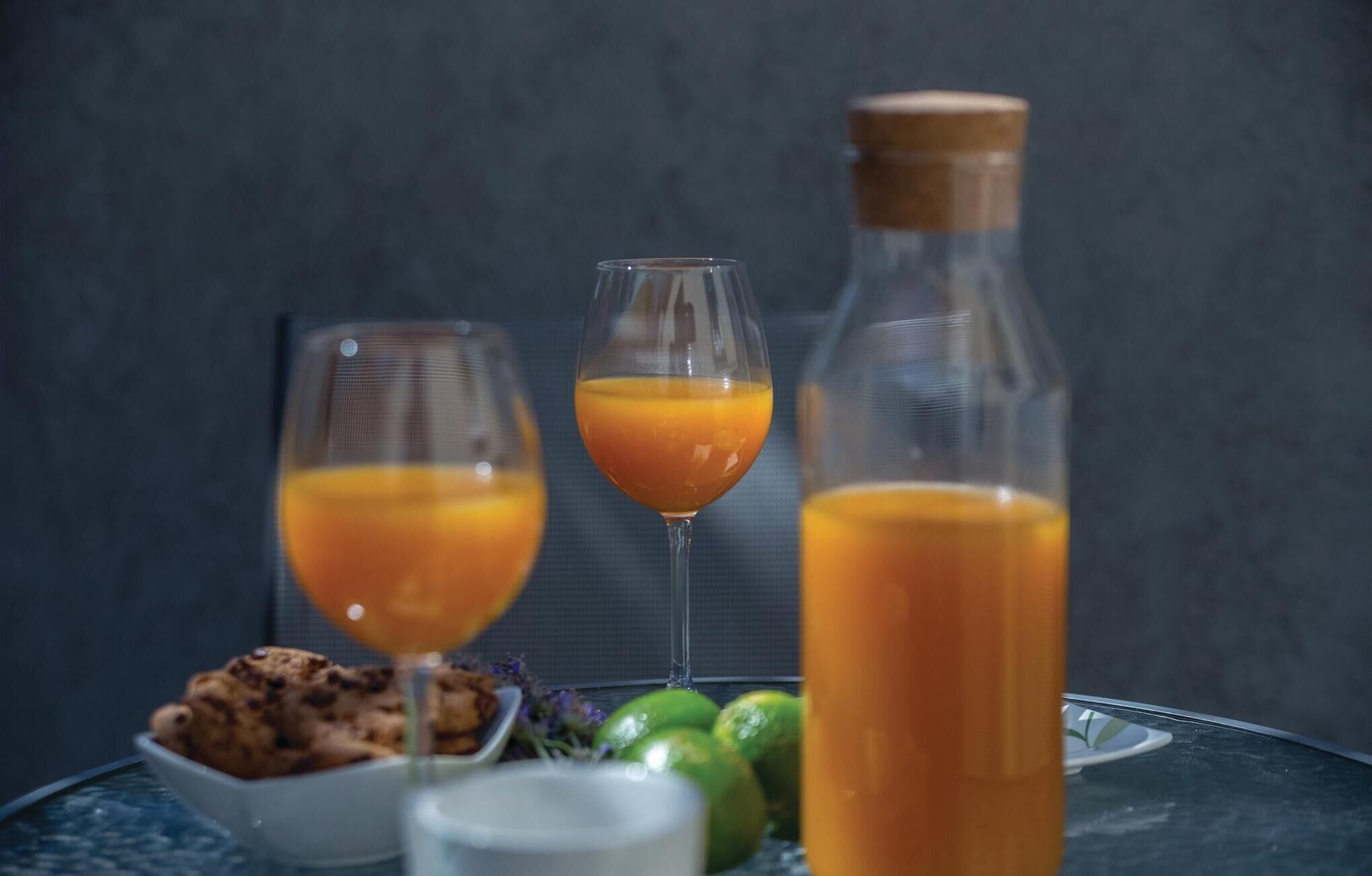 A table with two glasses of orange juice, a bottle of orange juice, a bowl of cookies, and some limes.
