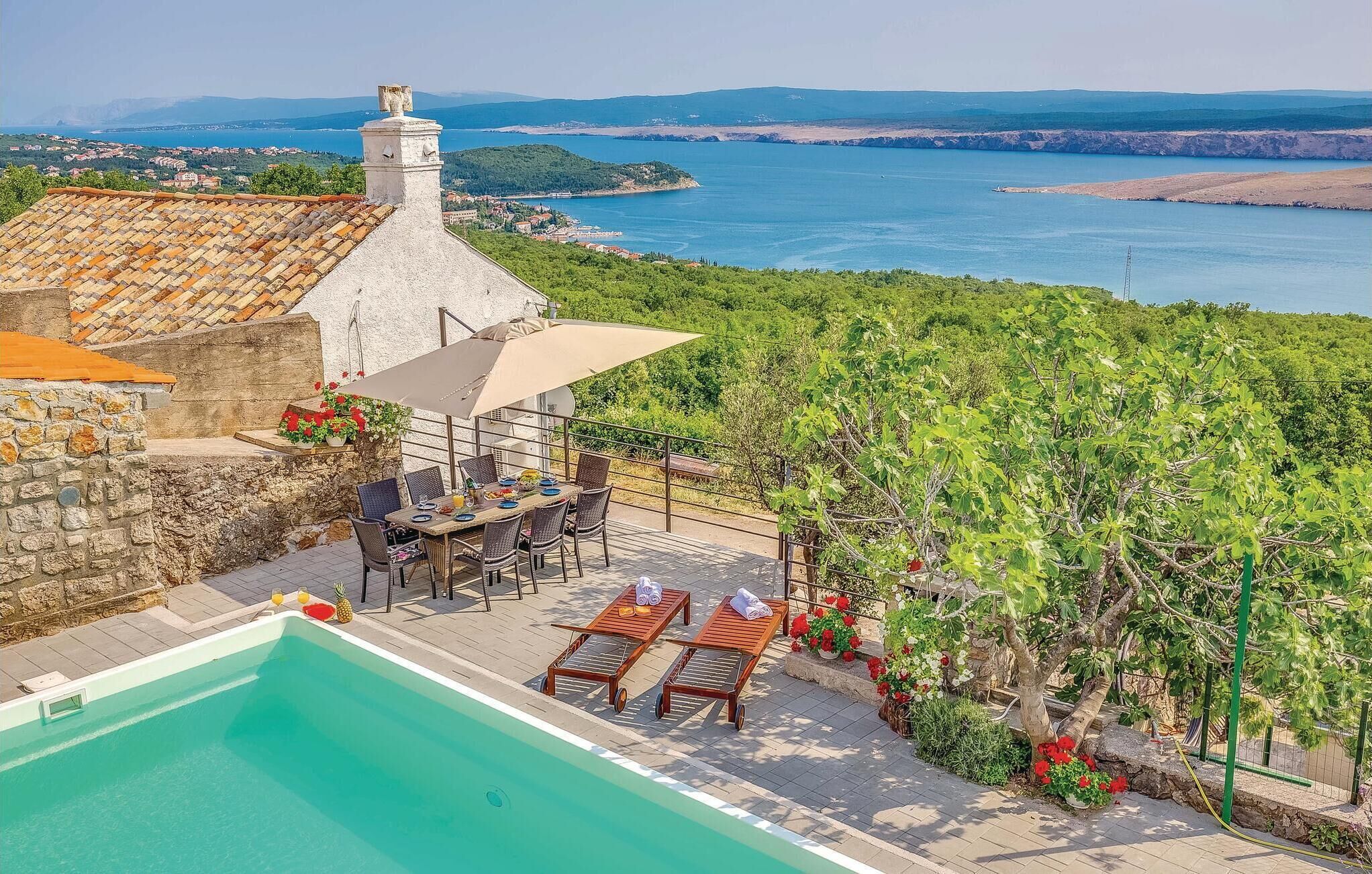 Un espace aménagé au bord de la piscine, avec une table et des chaises, une pergola abritant une table, et une vue sur la mer et les collines en arrière-plan.