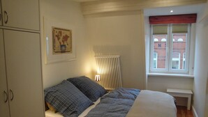 Une chambre à coucher comprenant un lit, un radiateur, une fenêtre avec des stores, un tableau accroché au mur et une armoire en bois.