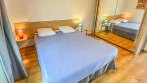 A neatly made bed with a blue bedspread and white pillows, a bedside table with a lamp, and a mirror reflecting the bed.