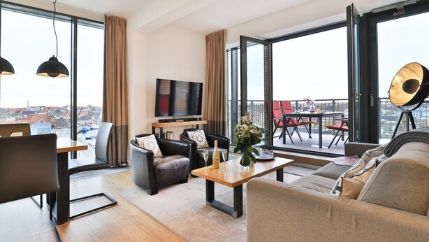 A modern living room with a sofa, armchair, and coffee table, a TV, and large windows with a city view. - Ohlerich storage app. 44 - Ohlerich Speicher App. 44 (Wismar)
