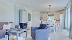 A modern living room with a sofa, coffee table, and a fireplace. There is a dining area with a table and chairs. The kitchen is visible in the background with white cabinets and a large window. - Aquamaritim beach house (Loddin)