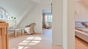 A spacious room with a high ceiling, a wooden floor, a bed, a wicker chair, and a small table with a vase. - Aquamaritim beach house (Loddin)