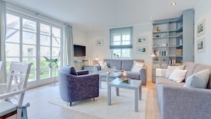 A modern living room with a grey sofa, a patterned armchair, and a coffee table. There is a bookshelf with decorative items and a television mounted on the wall. - Aquamaritim beach house (Loddin)