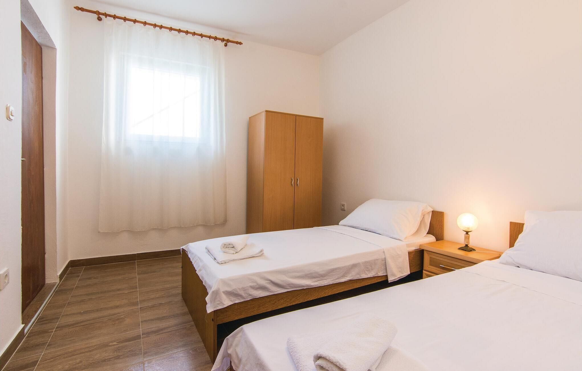 Quarto com duas camas de solteiro, guarda-roupa de madeira, mesa de cabeceira com abajur e janela com cortinas leves.