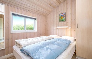 Ett sovrum med träväggar, ett stort fönster, en säng med blått och vitt sänglinne och en tavla på väggen.