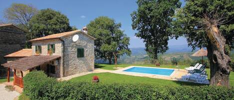 A stone house with a swimming pool, surrounded by well-maintained hedges and trees.